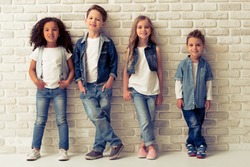 Full length portrait of cute little kids in stylish jeans clothes looking at camera and smiling, standing against white brick wall