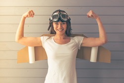 Beautiful young woman dressed like a pilot with toy wings is showing her muscles, smiling and looking upward, against gray wall