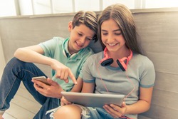 Teenage boy and girl with headphones are using gadgets, talking and smiling while sitting on the floor