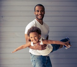 Handsome young Afro American father and his cute child are looking at camera and smiling while playing together. Kid is imitating an airplane