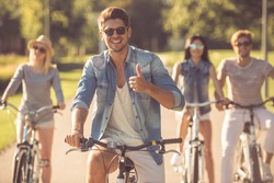 Handsome stylish guy is showing Ok sign, looking at camera and smiling while cycling with his friends in the park
