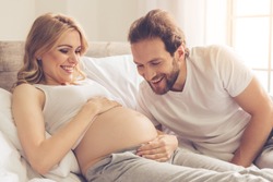 Beautiful pregnant woman and her handsome husband are smiling while spending time together in bed. Man is listening to baby in belly