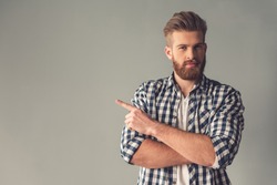 Handsome bearded man in casual clothes is pointing away, looking at camera and smiling, on gray background