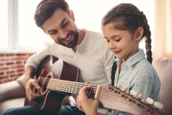Cute little girl and her handsome father are playing guitar and smiling while sitting on couch at home