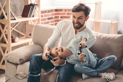 Cute little girl and her handsome father are playing game console and laughing while sitting on couch at home