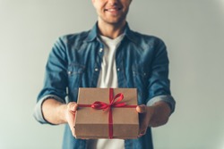Cropped image of handsome romantic guy smiling while holding a present for his couple, on gray background