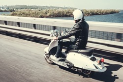 Guy in leather jacket and helmet is riding on scooter through the city bridge