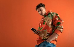 Fashion studio portrait of a young man in hoodie with smartphone posing over orange background.
