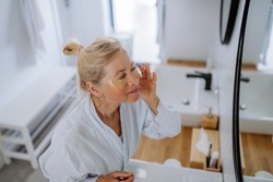 Beautiful senior woman in bathrobe, applying eye patches for puffiness while looking in the mirror