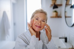 Beautiful senior woman in bathrobe, applying eye patches for puffiness while looking at camera