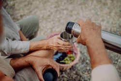 Close-up of senior couple having picnic at beach during autumn day, drinking hot tea from thermos.
