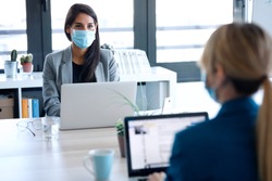 Shot of two business women wearing a hygienic face mask while work with laptops in the coworking space. Social distancing concept.