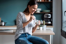 Shot of smiling young woman eating yogurt while sitting on stool in the kitchen at home.