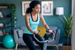 Shot of sporty african young woman exercising on smart stationary bike and listening to music at home.