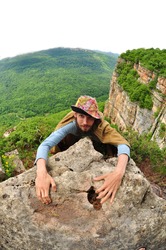 A man in a hat and a beard climbs on a ledge of rock, behind him a steep cliff and far away mountains covered with forests
