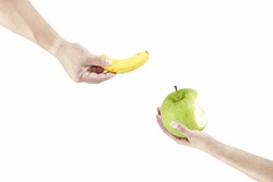 A hand of a man holding a yellow banana offering to a woman hand holding a green apple that is half eaten, isolated against white.