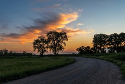Trees in silhouette against blue evening sky with orange tinted clouds and a gravel road on a summers day in rural Minnesota, United States
