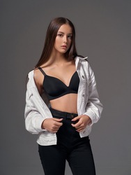 studio portrait of young slim tanned caucasian girl in black jeans, bra and white jeans jacket standing and posing against grey studio background