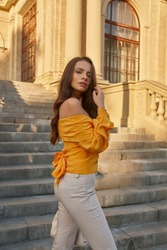 Young beautiful caucasian woman in beige pants and orange top holding handbag and posing at stairway. Pretty fashionable girl walking in the city on a summer evening