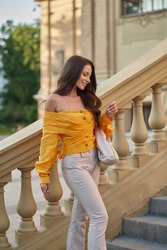 Young beautiful caucasian woman in beige pants and orange top holding handbag and posing at stairway. Pretty fashionable girl walking in the city on a summer evening