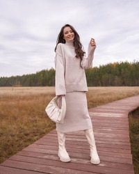 Smiling brunette woman in warm oversize sweater and skirt holds white handbag standing on wooden footbridge at countryside in autumn