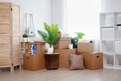moving day and relocation concept - stack of cardboard boxes, houseplants and other domestic things in living room in new house