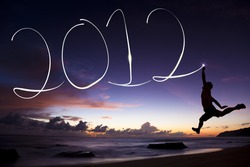 happy new year 2012. young man jumping and drawing 2012 by flashlight in the air on the beach before sunrise