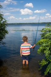 child is fishing in river on sunny summer day. boy casts a fishing rod, stands in water with his back to camera, a view of river, clouds in blue sky. Nature. Outdoor. Happy and interesting childhood