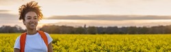Panorama header beautiful biracial African American girl teenager female young woman smiling and happy hiking with back pack in a field of yellow flowers at sunset or sunrise panoramic web banner.