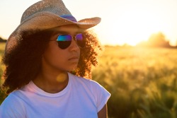 Beautiful happy mixed race African American female girl teenager young woman wearing reflective aviator sunglasses and cowboy hat in a cornfield at golden sunset or sunrise