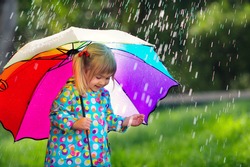 Funny cute toddler girl wearing waterproof coat with colorful umbrella playing in the garden by rainy and sunny day