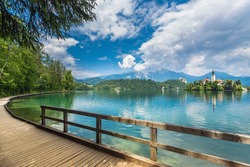 Wooden walkway around the lake Bled and the Catholic church of St. Mary situated on an island on Bled lake with mountains and villages on the background 