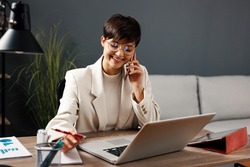 Young business woman working at home with laptop and papers on desk and talking on the phone