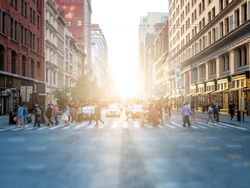 Crowd of anonymous people crossing the street at a busy intersection in Manhattan, New York City with the bright glow of sunset in the background