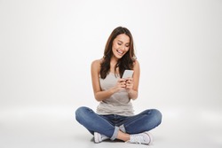 Happy brunette woman sitting on the floor and writing message on smartphone over gray background