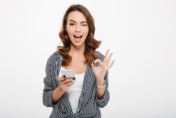 Portrait of a satisfied casual girl holding mobile phone and showing ok gesture isolated over white background