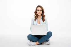 Portrait of a smiling casual girl holding laptop computer while sitting on a floor isolated over white background