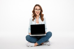 Portrait of a pretty casual girl showing blank screen laptop computer while sitting on a floor isolated over white background