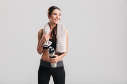 Healthy beautiful sportswoman wearing tracksuit with towel looking aside and holding bottle with fresh water isolated over gray background
