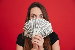Close up portrait of a young girl holding bunch of money banknotes at her face isolated over pink background