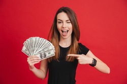 Portrait of a happy girl pointing finger at bunch of money banknotes and winking isolated over pink background