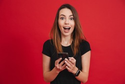 Photo of satisfied woman in black clothing looking on camera while using cell phone with joy over red background