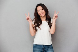 Portrait of asian attractive woman in casual t-shirt and jeans smiling and showing victory sign with both hands isolated over gray background