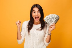 Portrait of an excited young woman holding money banknotes and celebrating isolated over yellow background