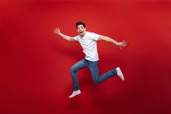 Full length portrait of an excited young man in white t-shirt jumping while celebrating success isolated over red background