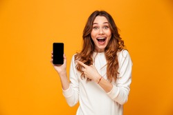 Surprised happy brunette woman in sweater showing blank smartphone screen and pointing on it while looking at the camera with open mouth over yellow background