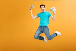 Portrait of a joyful young man in t-shirt holding bunch of money banknotes and celebrating while jumping isolated over yellow background