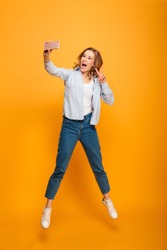 Full length portrait of young attractive woman smiling and showing victory sign while taking selfie with cell phone isolated over yellow background