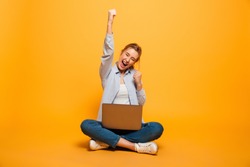 Portrait of a cheerful young girl with braces sitting on a floor with a laptop computer and celebrating success isolated over yellow background