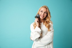 Portrait of a lovely young blonde woman in sweater showing credit card and looking away at copy space isolated over blue background